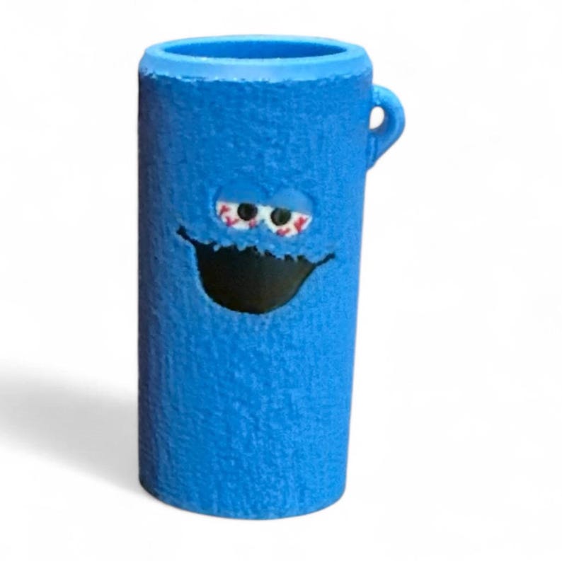 May include: A blue cylindrical mug with a cartoon face design. The mug features a textured surface, a small handle, and a wide open mouth. The eyes are red and white, and the mouth is black.