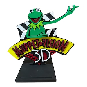 Könnte beinhalten: Bunte Kermit der Frosch-Figur mit den Worten "Muppet Vision 3D" in Rot, Gelb und Weiß. Die Figur steht auf einem schwarzen Sockel und zeigt eine Filmklappe. Der Frosch ist grün und lächelt breit.