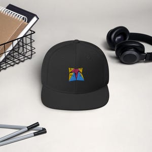 May include: Black flat-brimmed baseball cap with a colorful embroidered design on the front. The design features a geometric pattern with yellow, red, and blue colors. The cap has a structured fit and a classic, casual style.
