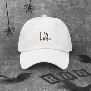 May include: White baseball cap with an embroidered design of two brooms and a vacuum cleaner. The cap is a classic six-panel design with a curved brim, perfect for Halloween or a witch-themed event.