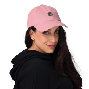 May include: A light pink baseball cap with a small, embroidered Rubik's Cube design on the front. The cap has a curved brim and a classic six-panel construction. The hat is made of a soft material.