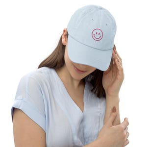 May include: A light blue baseball cap with a curved brim and a pink embroidered smiley face on the front. The cap is made of a soft material and has an adjustable strap. The hat is a casual accessory.