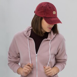 May include: A maroon baseball cap with a small embroidered house logo. The cap has a curved peak and an adjustable strap. The person is wearing a pink zip-up hoodie. The hoodie has a white drawstring.