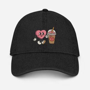 Könnte beinhalten: Schwarze Denim-Baseballkappe mit einem gestickten Design eines rosa Herzens mit Augen und einem Milchshake. Das Herz hält einen Strohhalm und der Milchshake hat einen rosa-weißen Wirbel mit einem rosa Herzen an der Seite.