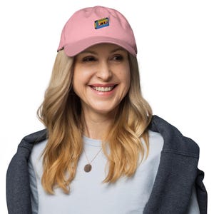 May include: A light pink baseball cap with a small embroidered cassette tape design in yellow, blue, and red. The cap has a curved brim and a classic six-panel design. The cap is worn by a person.
