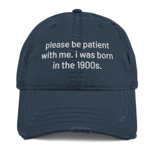 May include: Distressed navy blue baseball cap with white embroidered text that reads "please be patient with me. i was born in the 1900s."