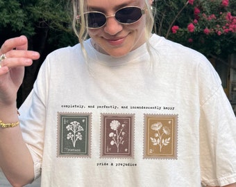 May include: A white t-shirt with a graphic of three vintage postage stamps featuring floral illustrations. The text "completely, and perfectly, and incandescently happy" is printed above the stamps. The text "pride & prejudice" is printed below the stamps.