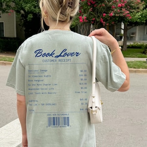 May include: Sage green t-shirt with the text "Book Lover" and a receipt design. The receipt lists fictional items like "Emotional Damage" and "3x Sleepless Nights." The shirt has a casual, relaxed fit.