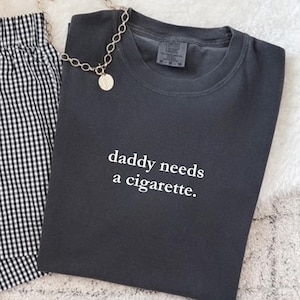 May include: A charcoal gray t-shirt with the text "daddy needs a cigarette" in white. The shirt is paired with black and white checkered shorts and a silver chain necklace with a round pendant.