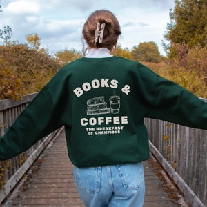 May include: A dark green sweatshirt with the text "BOOKS & COFFEE THE BREAKFAST OF CHAMPIONS" printed on the back. The text is in a white font and is accompanied by a graphic of a stack of books and a cup of coffee.