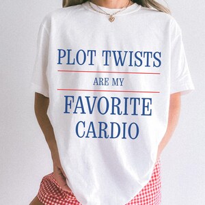 May include: White t-shirt with the text "PLOT TWISTS ARE MY FAVORITE CARDIO" in blue, with red lines separating the phrases. The shirt is a casual style, suitable for everyday wear.