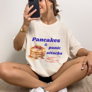 May include: A white t-shirt with a graphic of a stack of pancakes and the text "Pancakes & panic attacks" in blue. The text "Buy one get one!" is in red.
