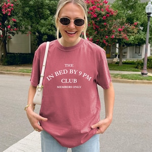 May include: A dusty rose-colored t-shirt with the text "THE IN BED BY 9 PM CLUB MEMBERS ONLY" in a white, curved font. The shirt is short-sleeved and appears to be made of a soft material. The person is wearing sunglasses and a white purse.