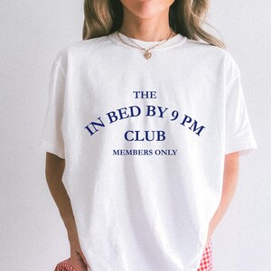 May include: White t-shirt with the text "THE IN BED BY 9 PM CLUB MEMBERS ONLY" in navy blue. The shirt has short sleeves and a crew neck. The overall style is casual and relaxed.