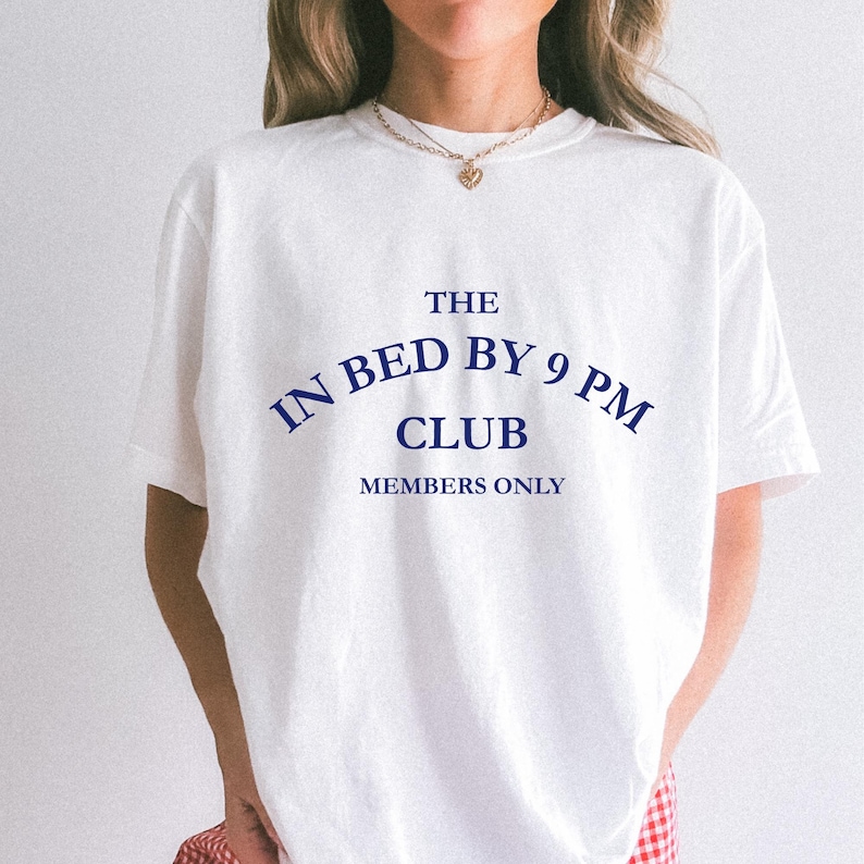 May include: White t-shirt with the text "THE IN BED BY 9 PM CLUB MEMBERS ONLY" in navy blue. The shirt has short sleeves and a crew neck. The overall style is casual and relaxed.