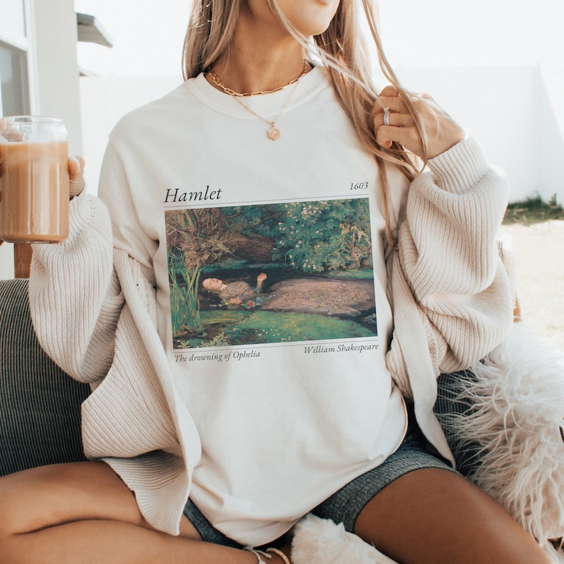 Op de afbeelding: Wit t-shirt met een afbeelding van een schilderij waarop een vrouw in een waterpartij ligt. Boven het schilderij staat de tekst "Hamlet 1603". Onder het schilderij staat de tekst "The drowning of Ophelia" en "William Shakespeare".