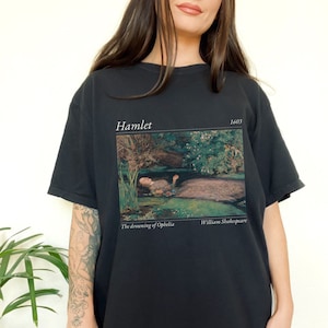 Op de afbeelding: Zwart T-shirt met een grafische print van een schilderij dat een vrouw in een rivier voorstelt. De tekst "Hamlet 1603" staat boven de afbeelding en "The drowning of Ophelia William Shakespeare" staat onder de afbeelding.