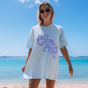 May include: A light blue t-shirt featuring a purple tiger graphic and the word "EASY" printed on the front. The model is wearing the shirt and standing on a beach.