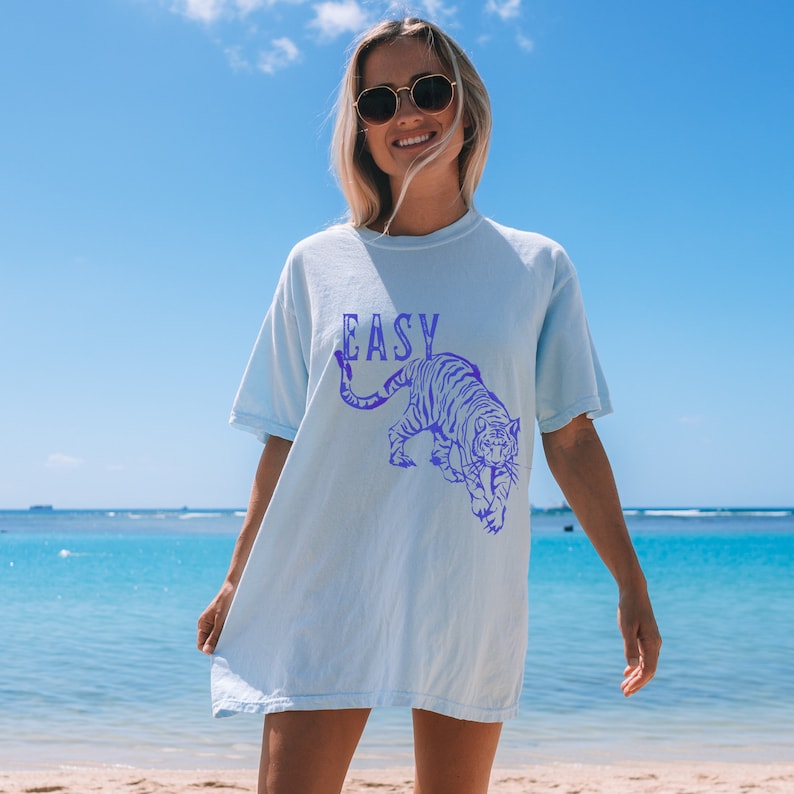 May include: A light blue t-shirt featuring a purple tiger graphic and the word "EASY" printed on the front. The model is wearing the shirt and standing on a beach.