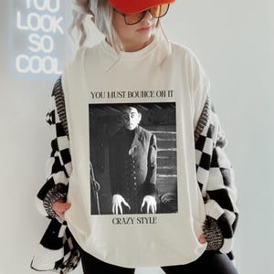 May include: White long-sleeve sweatshirt with a black and white checkered pattern on the sleeves. The front of the sweatshirt features a black and white image of a man with the text "You must bounce on it" and "Crazy Style" printed below the image.