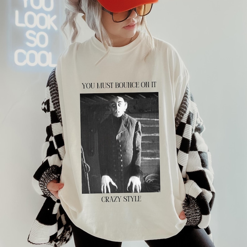 May include: White long-sleeve sweatshirt with a black and white checkered pattern on the sleeves. The front of the sweatshirt features a black and white image of a man with the text "You must bounce on it" and "Crazy Style" printed below the image.