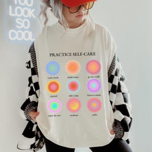 May include: White t-shirt with the text "PRACTICE SELF-CARE" and nine colorful circles with text describing self-care activities, such as "read a book", "drink water", "go on a walk", "journal", "take a nap", "listen to music", "enjoy the sun", "meditate", and "smile".