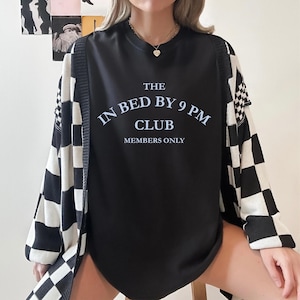 May include: Black crew neck sweatshirt with the text "THE IN BED BY 9 PM CLUB MEMBERS ONLY" in white. The sweatshirt is paired with a black and white checkered cardigan. A gold necklace with a heart pendant is visible.