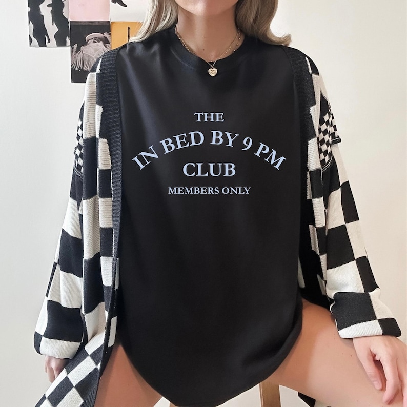 May include: Black crew neck sweatshirt with the text "THE IN BED BY 9 PM CLUB MEMBERS ONLY" in white. The sweatshirt is paired with a black and white checkered cardigan. A gold necklace with a heart pendant is visible.