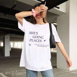 Puede incluir: Camiseta blanca con el texto "She's Going Places (I'm She and It's to Get Another Book)" en letras azul marino con dos peque&ntilde;as estrellas a cada lado del texto.