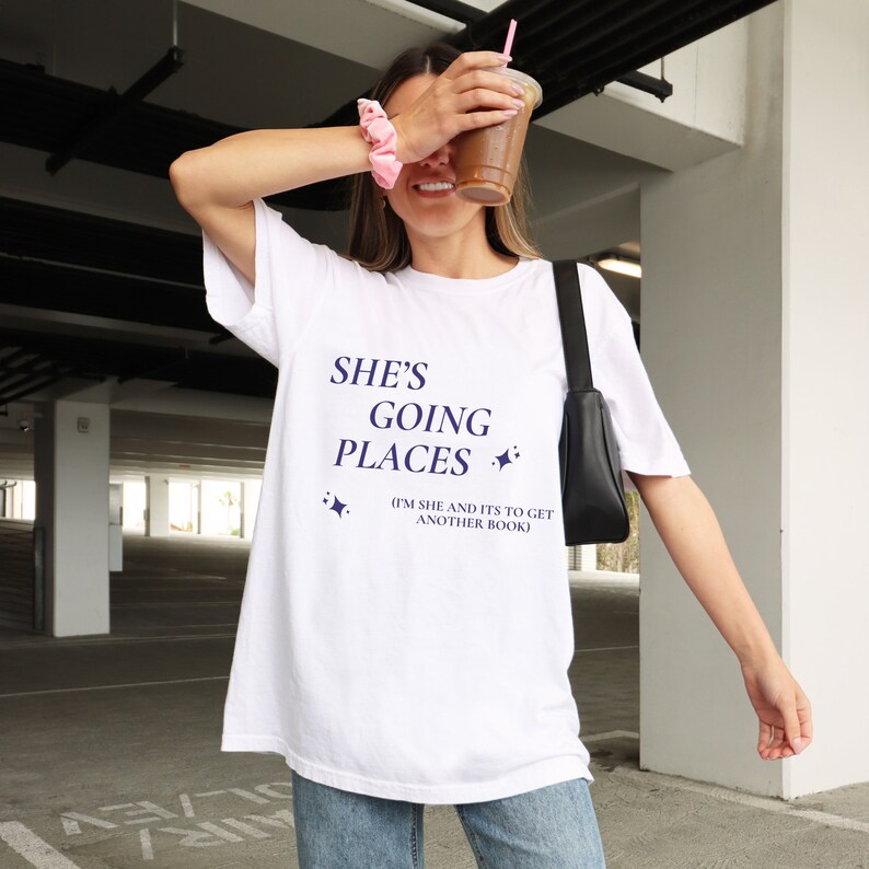 Puede incluir: Camiseta blanca con el texto "She's Going Places (I'm She and It's to Get Another Book)" en letras azul marino con dos peque&ntilde;as estrellas a cada lado del texto.