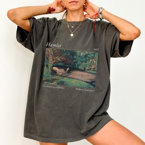 Op de afbeelding: Een oversized donkergrijs T-shirt met een print van een schilderij dat een vrouw in een rivier laat zien. De tekst "Hamlet" en "1603" staat boven het schilderij gedrukt. De tekst "The drowning of Ophelia" en "William Shakespeare" staat onder het schilderij gedrukt.
