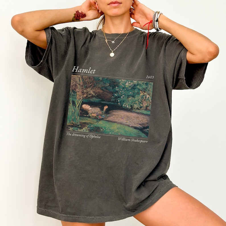 Op de afbeelding: Een oversized donkergrijs T-shirt met een print van een schilderij dat een vrouw in een rivier laat zien. De tekst "Hamlet" en "1603" staat boven het schilderij gedrukt. De tekst "The drowning of Ophelia" en "William Shakespeare" staat onder het schilderij gedrukt.