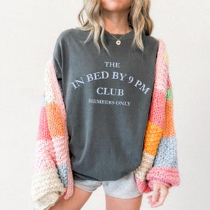 May include: A dark gray t-shirt with the text "THE IN BED BY 9 PM CLUB MEMBERS ONLY" in white. The person is wearing a colorful patchwork cardigan with pink, orange, and cream blocks. The person is also wearing gray shorts.