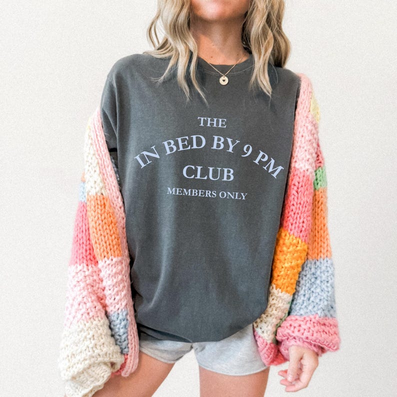 May include: A dark gray t-shirt with the text "THE IN BED BY 9 PM CLUB MEMBERS ONLY" in white. The person is wearing a colorful patchwork cardigan with pink, orange, and cream blocks. The person is also wearing gray shorts.