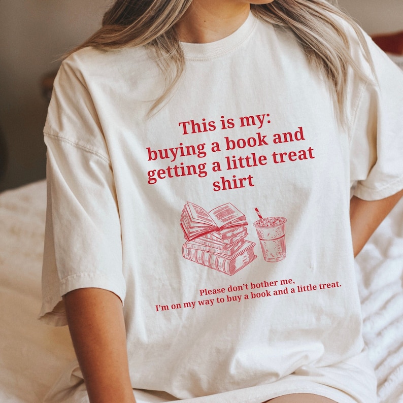 May include: A white t-shirt with red text that reads "This is my: buying a book and getting a little treat shirt". The text is accompanied by an illustration of a stack of books and a cup of iced coffee.