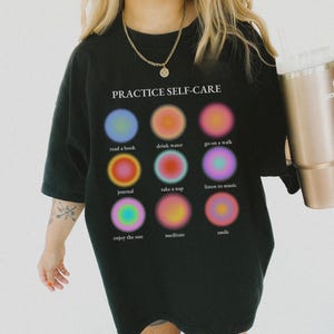 May include: Black t-shirt with the text "PRACTICE SELF-CARE" and a grid of nine colorful circles, each with a different self-care activity listed below it. The activities are: read a book, drink water, go on a walk, journal, take a nap, listen to music, enjoy the sun, meditate, and smile.