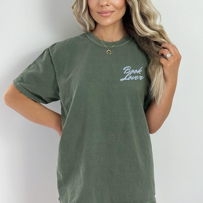 May include: Olive green t-shirt with the text "Book Lover" in a light blue script. The shirt has a crew neck and short sleeves. The fabric appears soft and comfortable.