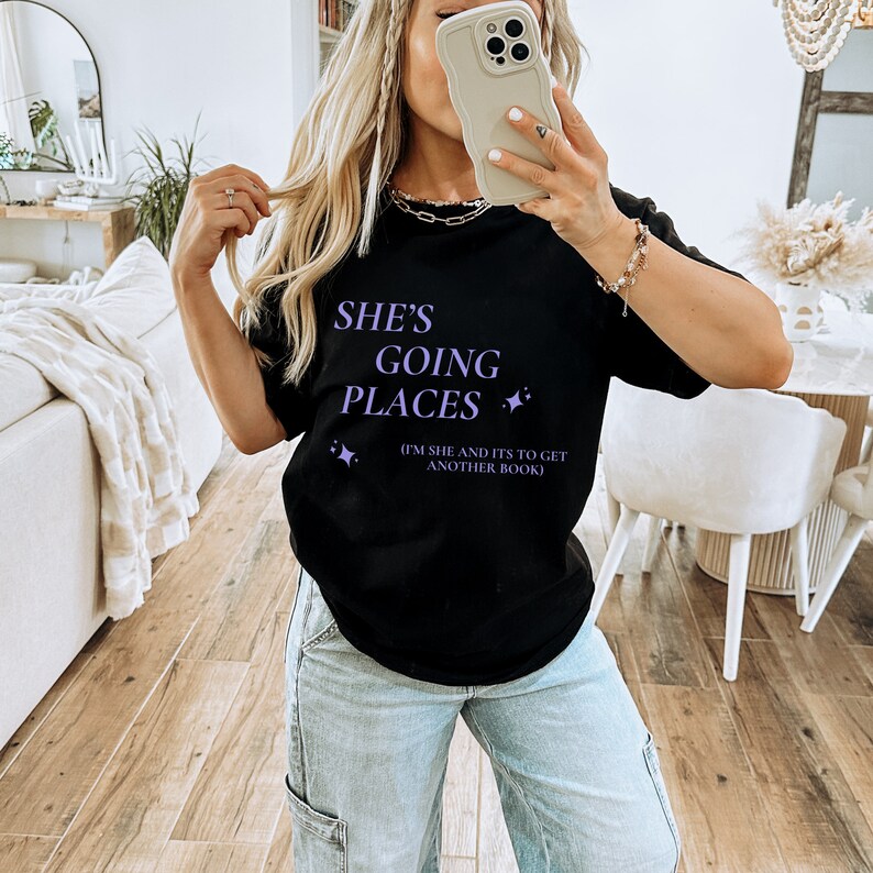 Puede incluir: Camiseta negra con el texto "She's Going Places (I'm She and It's to Get Another Book)" en letras moradas con peque&ntilde;as estrellas moradas.