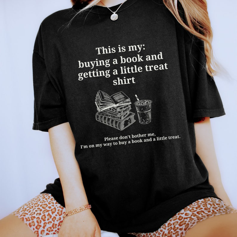 May include: Black t-shirt with white text that reads "This is my: buying a book and getting a little treat shirt. Please don't bother me, I'm on my way to buy a book and a little treat."
