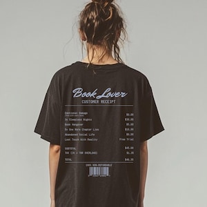 May include: Black t-shirt with the text "Book Lover" and a customer receipt design printed on the back. The receipt lists fictional items like "Emotional Damage" and "Sleepless Nights". The shirt is a dark color and has a casual style.