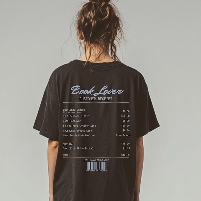 May include: Black t-shirt with the text "Book Lover" and a customer receipt design printed on the back. The receipt lists fictional items like "Emotional Damage" and "Sleepless Nights". The shirt is a dark color and has a casual style.