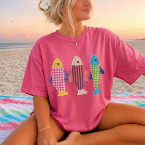 May include: A pink short-sleeved t-shirt featuring three fish appliqués on the front. The left fish has a pink and white gingham pattern, the middle fish has purple and orange vertical stripes, and the right fish has a yellow and green plaid pattern.