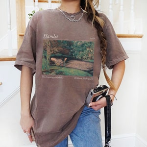 Op de afbeelding: Een oversized bruin t-shirt met een vintage print van een schilderij dat een vrouw in een rivier laat zien. De print bevat de tekst "Hamlet 1603", "The drowning of Ophelia" en "William Shakespeare".