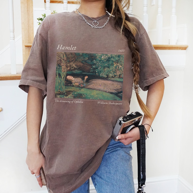 Op de afbeelding: Een oversized bruin t-shirt met een vintage print van een schilderij dat een vrouw in een rivier laat zien. De print bevat de tekst "Hamlet 1603", "The drowning of Ophelia" en "William Shakespeare".