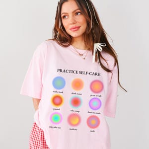 May include: Pink t-shirt with the text "PRACTICE SELF-CARE" and a graphic of nine colorful circles with self-care activities listed below each circle. The activities are: read a book, drink water, go on a walk, journal, take a nap, listen to music, enjoy the sun, meditate, and smile.