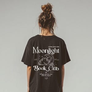May include: Black t-shirt with the text "COME JOIN THE Moonlight Book Club" in white. The design includes a crescent moon, clouds, and stars. The shirt is a casual, oversized fit.