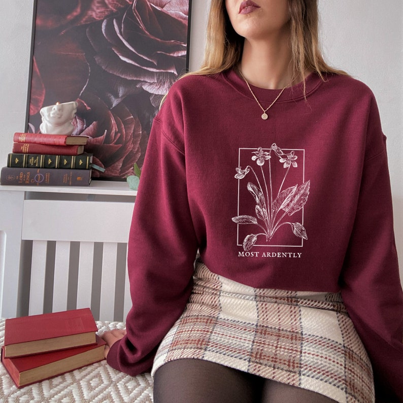 May include: A maroon sweatshirt with a white floral design and the text "MOST ARDENTLY" printed on the front. The sweatshirt is worn by a person wearing a plaid skirt.