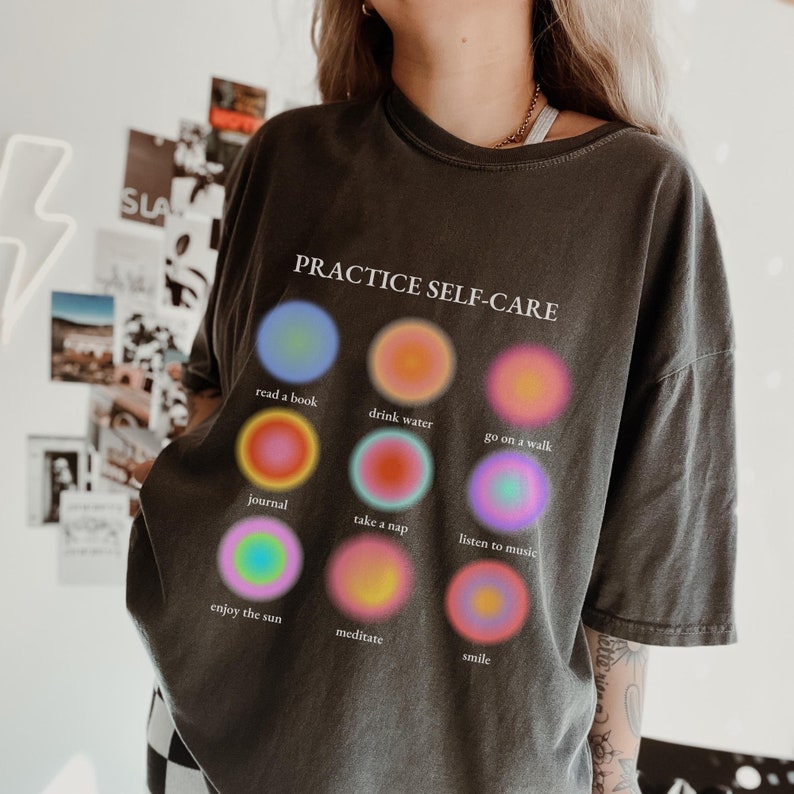 May include: A black t-shirt with the text "PRACTICE SELF-CARE" and a grid of nine colorful circles. Each circle has a different self-care activity written below it, including "read a book", "drink water", "go on a walk", "journal", "take a nap", "listen to music", "enjoy the sun", "meditate", and "smile".