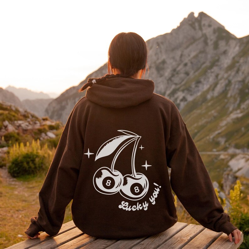 Op de afbeelding: Bruine hoodie met een witte afbeelding van twee kersen met het nummer 8 op elk. De kersen zitten aan een steel met bladeren en de tekst "Lucky you!" staat eronder.