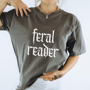 May include: A dark gray t-shirt with the words "feral reader" in a gothic font. The shirt has short sleeves and a crew neck. The fabric appears soft and slightly faded.